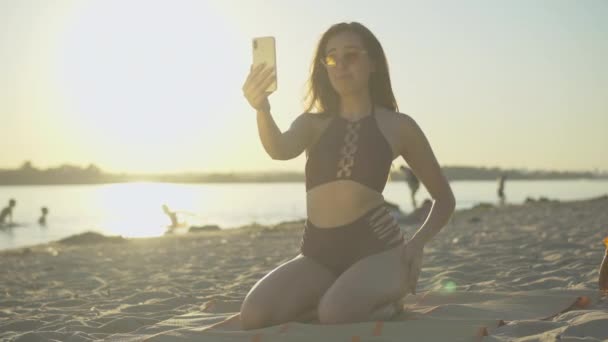 Mujer delgada linda alegre hablando en la cámara selfie en el teléfono inteligente como amigo alegre unirse a ella en la playa de arena. Retrato de dos turistas caucásicos positivos usando chat al atardecer en un resort de verano. — Vídeo de stock