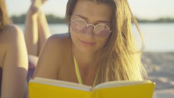 Foto close-up dari wanita muda yang cantik membaca buku saat matahari terbenam di pantai berpasir. Wanita Kaukasia dengan kacamata hitam yang menawan menikmati hobi di resor musim panas di bawah sinar matahari. Pariwisata dan gaya hidup. — Stok Video
