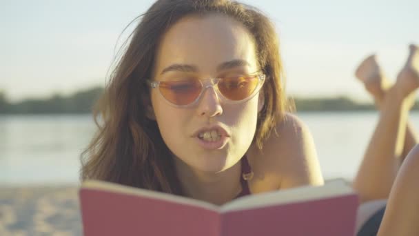Close-up face of charming brunette young woman reading out loud. Portrait of confident beautiful Caucasian tourist in sunglasses resting at sunset on sandy summer beach. Cute lady enjoying hobby. — Stock Video