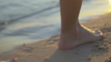 Gün batımında kumlu sahil boyunca yürüyen aşırı yakın plan beyaz kadın ayakları. Gün batımında denizde ya da okyanus kıyısında gezinen tanınmayacak kadar zayıf ve çıplak ayaklı bir kadın. Doğada güzellik, eğlence, turizm.