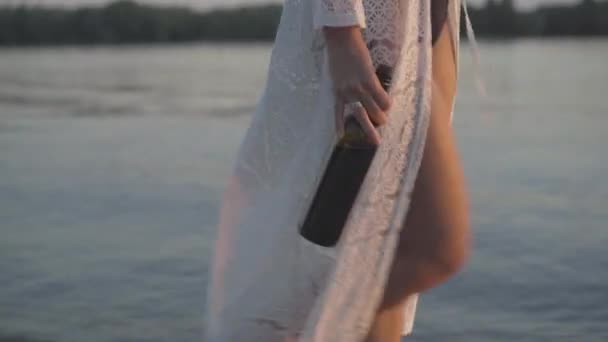 Femme caucasienne bronzée et méconnaissable en maillot de bain et en robe marchant avec une bouteille de vin le long de la rivière ou de la mer. Slim jeune touriste femelle se promener sur la plage au coucher du soleil avec boisson. — Video