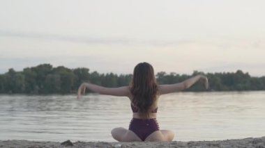 Nilüfer çiçekli sahilde poz veren zayıf, kendine güvenen dişi yogi 'nin geniş açılı görüntüsü. Esmer, beyaz bir kadın gün batımında yoga yapıyor. Sağlıklı yaşam tarzı, yaşam dengesi