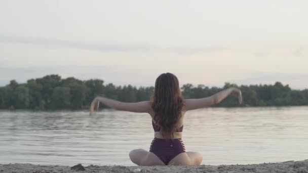 Retrovisore ampio colpo di sottile fiducioso yogi femminile seduto in posa loto sulla spiaggia sabbiosa sulla riva del fiume. Bruna giovane donna caucasica che pratica yoga all'aperto al tramonto. Stile di vita sano, equilibrio di vita — Video Stock