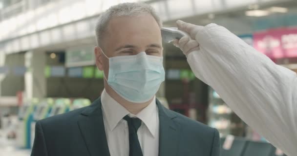 Närbild porträtt av mellanvuxen affärsman i ansiktsmask står i flygplatsen avgångsområde som anställd mäter sin temperatur med kontaktlös termometer, och lämnar. Bio 4k ProRes HQ. — Stockvideo