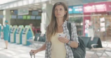 Havaalanında akıllı telefon kullanan ve kalkış panosuna bakan güzel beyaz bir kadının portresi. Kadın turist içeride uçmayı bekliyor. Turizm ve seyahat. Sinema 4k ProRes Merkezi.