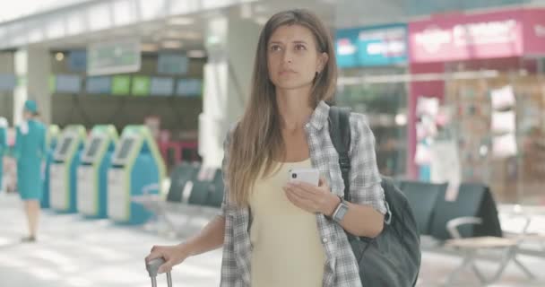 Portrait of beautiful young Caucasian woman using smartphone in airport and looking at departure board. Female tourist waiting for flight indoors. Tourism and travelling. Cinema 4k ProRes HQ. — Stock Video