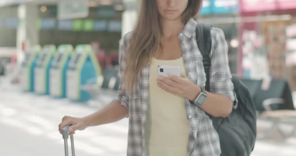 Невідома молода жінка стоїть з багажем у зоні відправлення аеропорту та перевіряє час польоту на смартфоні. Сучасні технології в туризмі. Кінотеатр 4k ProRes HQ . — стокове відео
