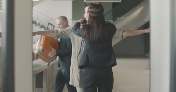 Vista trasera del empleado del aeropuerto examinando a una mujer caucásica de mediana edad con anteojos en las puertas de seguridad como hombre que se va con el equipaje en el fondo. Inspección física en aeropuerto. Sede del cine 4k ProRes. — Vídeos de Stock