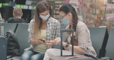 Beyaz maskeli anne ve kız akıllı telefondan kalkış zamanını kontrol ediyor. Havaalanındaki bekleme salonunda oturan kadın turistlerin portresi. Covid-19 'da turizm. Sinema 4k ProRes Merkezi.