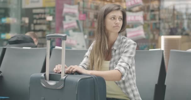 Egy fiatal, unatkozó kaukázusi nő portréja, aki csomagokkal a váróban ül. A barnahajú turista várja az indulást a repülőtéren, vasútállomáson vagy buszpályaudvaron. Idegenforgalmi koncepció. Cinema 4k ProRes főhadiszállás. — Stock videók
