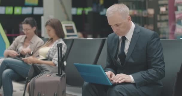 Серьезный кавказский бизнесмен среднего возраста, пользующийся ноутбуком в зале ожидания аэропорта с людьми, разговаривающими на заднем плане. Уверенный человек ждет отъезда на железнодорожной или автобусной станции. Штаб-квартира кинотеатра 4k ProRes. — стоковое видео