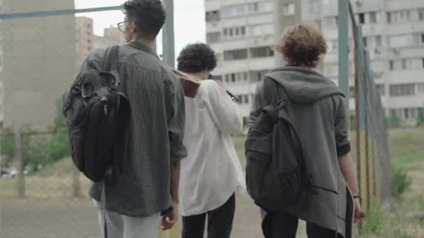 Back view of three young boys dressed in 1980s style entering outdoor sport court. Camera follows cheerful multiethnic fellows with backpacks and guitar. Retro and vintage concept. — Stock Video