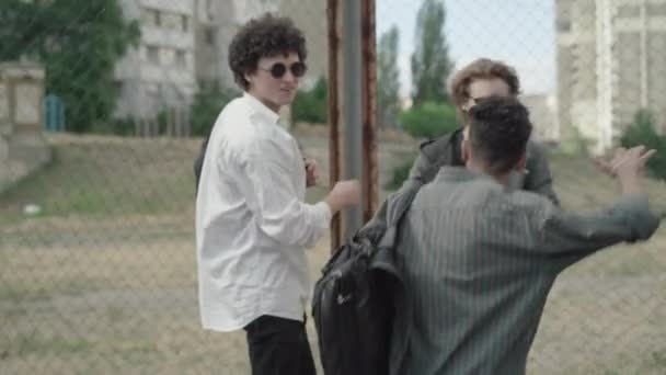 African American high-school student greeting Caucasian friends outdoors in 1980s. Camera follows joyful young man making fake fight with fellows at mesh fence and laughing. Male friendship. — Stock Video