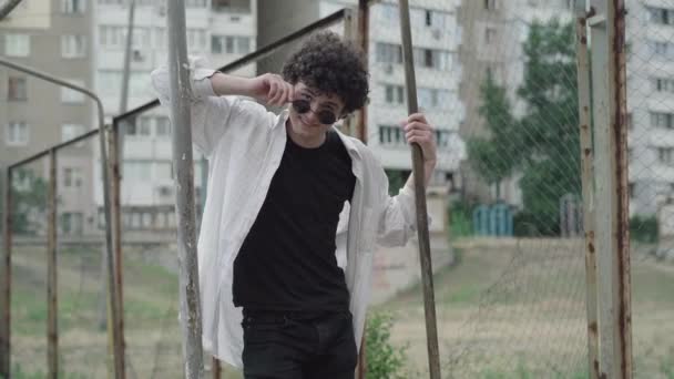 Retrato de un joven caucásico con estilo en la década de 1980 o 1990 ropa poner gafas de sol y guiño a la cámara. Medio tiro de confianza rizado chico coqueteando al aire libre en el centro de la ciudad. — Vídeo de stock