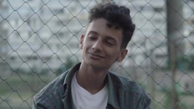 Kıvırcık saçlı, kahverengi gözlü ve kameraya bakıp gülümseyen yakışıklı Afro-Amerikan adamın portresi. Akşam karanlığında şehir merkezinde poz veren pozitif genç bir adam. Göçmen çitte oturuyor..