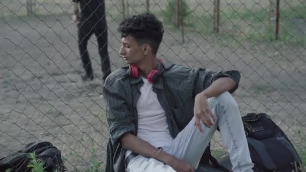 Портрет упевненого афроамериканського підлітка, який дивиться рюкзак, залишений непізнаним хлопцем і слухаючи музику в навушниках. Самотній хлопець сидить на самоті, коли люди грають у футбол.. — стокове відео