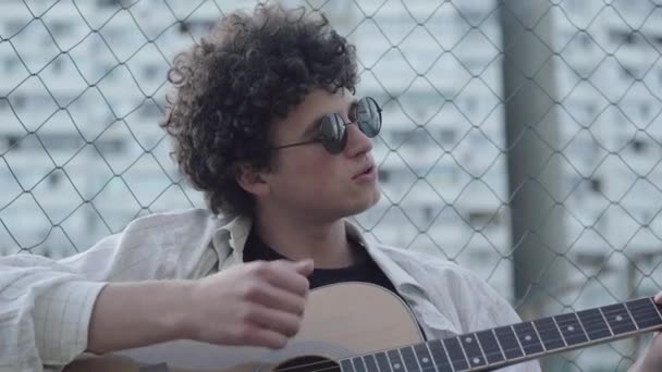 Романтичний молодий кавказький чоловік у сонцезахисних окулярах грає на гітарі в сутінках і співає. Портрет красивого кучерявого хлопця, який насолоджується хобі в 1990-х або 1980-х роках. Довірливий музикант відпочиває надворі.. — стокове відео