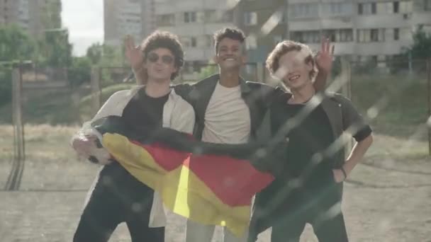 Tres felices amigos multiétnicos posando con pelota deportiva y bandera alemana detrás de una valla de malla. Retrato de jóvenes alegres celebrando la reunión de Alemania Oriental y Occidental en 1989. Hito histórico. — Vídeos de Stock