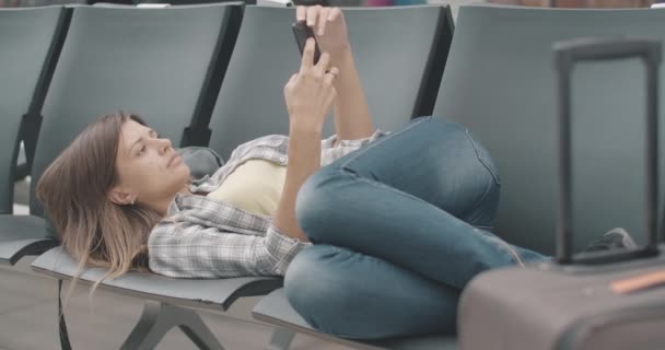 Mujer joven aburrida y cansada tumbada en sillas en la sala de espera del aeropuerto y usando un teléfono inteligente. Retrato de morena hermosa turista caucásica esperar a la salida retardada. Sede del cine 4k ProRes. — Vídeos de Stock