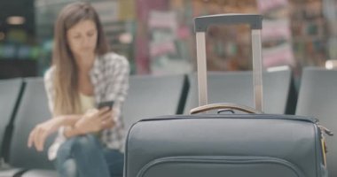 Bekleme salonunda duran seyahat çantasının yakın plan fotoğrafı. Akıllı telefonlu, bulanık, beyaz bir kadın bavulunu alıp bavulunu alıp gidiyor. Seyahat eden bir bayan turist. Sinema 4k ProRes Merkezi.