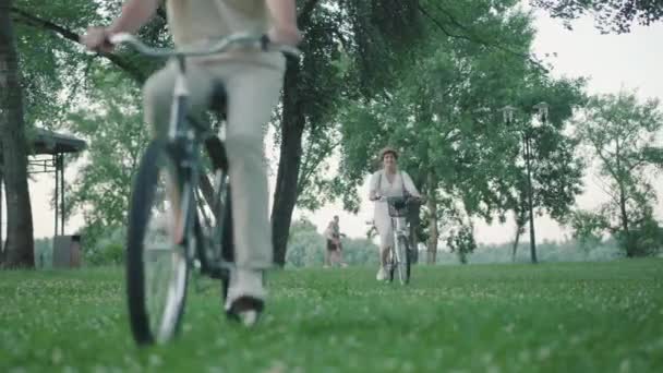 Positivo mediados de adulto hombre y mujer montar en bicicleta en el césped verde en el parque de verano y salir. Feliz sonrisa elegante pareja caucásica disfrutando de hobby al aire libre en el día soleado. Ocio y diversión. — Vídeo de stock