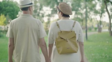 Rahatlamış yetişkin çiftlerin yaz parkında gezintilerinin arka görüntüsü. Güneşli bir günde, beyaz karı ve koca el ele tutuşup sokakta yürürken mutlu olun. Sevgi dolu erkek ve kadın dışarıda dinleniyor..