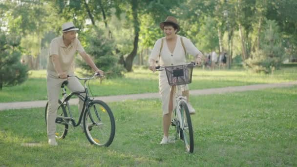 Πορτρέτο του χαρούμενου μεσήλικα Καυκάσια ζευγάρι ιππασία ποδήλατα στο ηλιόλουστο καλοκαιρινό πάρκο. Σύζυγος ακολουθεί αξιολάτρευτη γυναίκα φεύγει με ποδήλατο. Ευτυχισμένη οικογένεια που απολαμβάνει το χόμπι της. Δέσμευση, αγάπη. — Αρχείο Βίντεο
