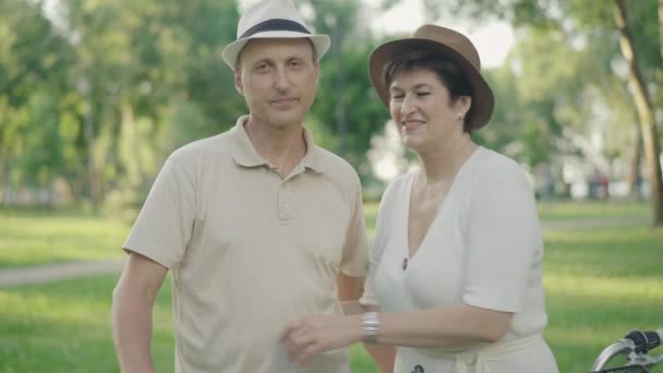 Счастливая любящая женщина обнимает мужа в парке и улыбается в камеру. Мужчина смотрит на жену с любовью. Портрет позитивной белой пожилой пары, наслаждающейся закатом в летнем парке. — стоковое видео