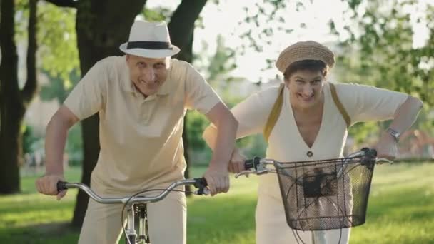 해질 녘 자전거를 타고 서 있는 긍정적 인 커플의 모습. 즐거운 성인 백인 남성과 여성이 카메라 앞에서 얼굴을 만들고 웃고 있습니다. 즐거운 가족들이 여름 공원에서 즐거운 시간을 보내다. — 비디오