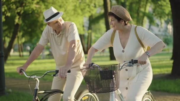 Pareja positiva sentada en bicicletas bajo los rayos del sol e imitando el paseo. Retrato de alegre hombre y mujer caucásicos de mediana edad divirtiéndose en la noche de verano al aire libre. Familia feliz descansando en el parque de verano. — Vídeos de Stock