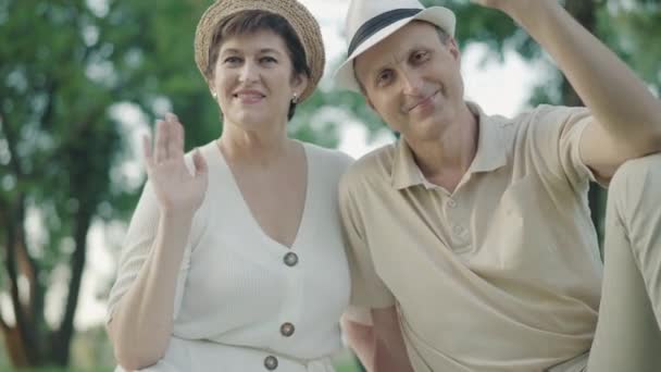 Portrét veselého středního dospělého páru mávající a usmívajícího se před kamerou. Šťastný kavkazský muž a žena pózují v letním parku za slunečného dne. Radostná rodina si užívá víkendy venku.