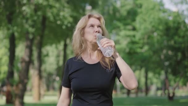 Camera nadert tot tevreden volwassen vrouw drinken verfrissend water uit fles in de zomer park. Portret van een zelfverzekerde blanke sportvrouw die buiten aan het trainen is. Levensstijl. — Stockvideo