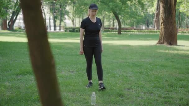 Amplio tiro de mujer caucásica delgada y segura en ropa deportiva negra y gorra atando zapatillas de deporte cordones, tomando botella de agua y caminando. Retrato de mujer deportiva de mediana edad haciendo ejercicio en el soleado parque de verano. — Vídeos de Stock