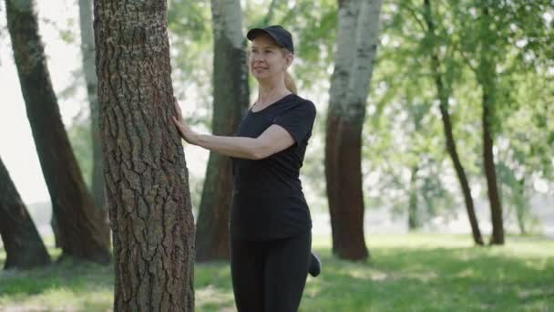 Confident smiling Caucasian sportswoman stretching legs in sunny park in the morning. Portrait of slim positive woman in sportswear and cap warming up before training outdoors. Sport concept. — Stock Video