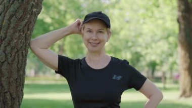 Siyah spor giyimli, şapkalı, güneşli yaz parkında kameraya gülümseyen orta yaşlı bir kadın. Eğitimden sonra dışarıda poz veren mutlu beyaz kadın. Spor, spor, spor..