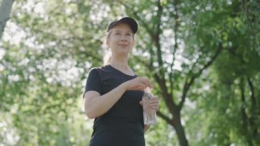 Açık havada güneş ışığında su şişesini açıp gülümseyen sporcu kadının portresi. Şapkalı ve spor kıyafetli zayıf, beyaz kadın yaz parkında güneş ışığında dikiliyor..