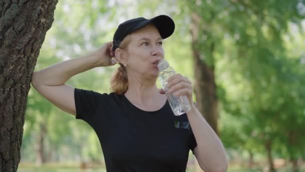 Mosolygó középkorú sportoló nő támaszkodik a fa a parkban és az ivóvíz. Portré pozitív kaukázusi nőről, aki friss levegőn pihen a szabadban. Sport, egészséges életmód. — Stock videók