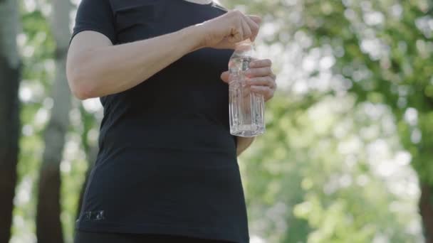 Felismerhetetlen középkorú nő sportruházatban felnyitja a frissítő vizet a nyári parkban. Sportos kaukázusi hölgy áll a szabadban a napfényben. Sportnő edzés a napos reggelen. — Stock videók