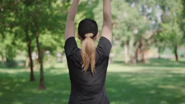 Visszapillantás a szőke középkorú, fekete sapkás és pólós sportos nőre, aki felemeli a kezét és hajlít. Vékony kaukázusi nő gyakorol a szabadban a napsütéses napon. Sportos női edzés a nyári parkban. — Stock videók