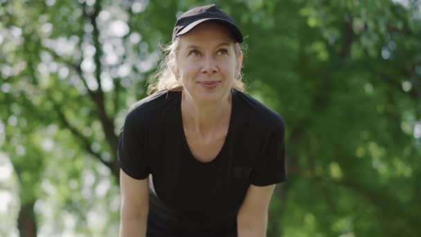 Actieve middenvolwassen vrouw rustend na de training in het zomerpark. Zelfverzekerde vermoeide blanke sportvrouw zucht en veegt het voorhoofd met de hand. Portret van een sportieve dame buiten. — Stockvideo