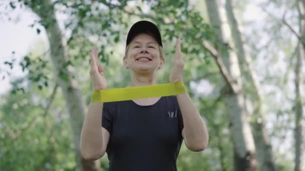 Happy mid-adult sportswoman training hands stretching resistance band. Portrait of cheerful Caucasian woman in cap and T-shirt exercising outdoors in sunny summer park. Training of confident lady. — Stock Video