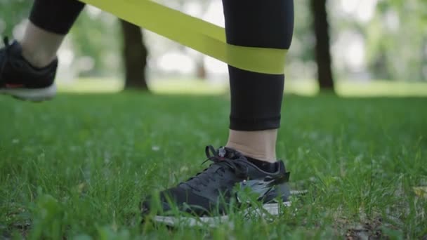 Ноги спортсменки середнього віку розтягують смугу опору на відкритому повітрі. Невпізнавана кавказька жінка в кросівках і тренувальних лапах спортивного одягу в сонячному літньому парку. Думка про здоровий спосіб життя. — стокове відео