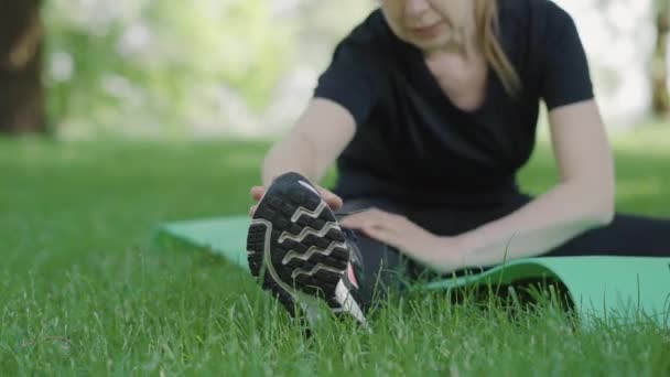 Yogui irreconocible de adulto medio calentándose antes de entrenar en el parque de verano. Mujer caucásica borrosa de mediana edad que se estira mientras está sentada en la esterilla de ejercicio. Centrado en zapatillas de deporte en la parte delantera. — Vídeos de Stock