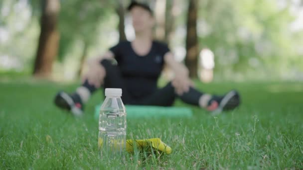 Eine Flasche erfrischendes Wasser steht auf dem Rasen im Sommerpark, im Hintergrund wärmt sich eine verschwommene kaukasische Sportlerin auf. Unerkennbare erwachsene Frau sitzt auf Yogamatte und dehnt sich. — Stockvideo