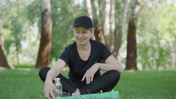 Θετική σπορ γυναίκα μυρωδιά και δάγκωμα μήλο, όπως κάθεται στο στρώμα άσκησης στο πάρκο το καλοκαίρι. Πορτρέτο με αυτοπεποίθηση Καυκάσιος θετικός γιόγκι ξεκουράζεται μετά την προπόνηση. — Αρχείο Βίντεο