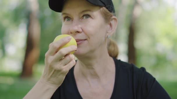 在夏天的公园里，穿着运动服，头戴帽子，头戴美味苹果的快乐白人女性的特写镜头。一个悠闲的中年女运动员在户外享受阳光的形象。健康的生活方式. — 图库视频影像