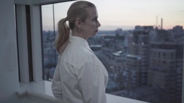 Side view of confident mid-adult businesswoman looking at evening city through office window. Portrait of beautiful serious Caucasian woman in white shirt thinking on balcony in business center. — Stock Video