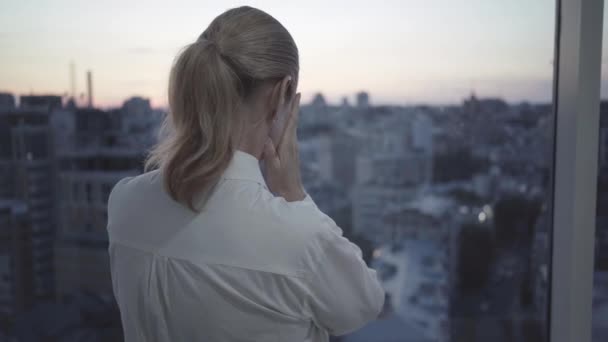 Depressed mid-adult businesswoman standing in front of window looking at city in the evening. Back view of stressed Caucasian woman standing on balcony and thinking at dusk. Overworking concept. — Stock Video