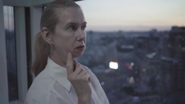 Detailní portrét přemýšlivé sebevědomé ženy stojící za soumraku na proskleném balkóně. Středně dospělá běloška tráví večer v kanceláři v městském městě. Řízení a přepracování. — Stock video