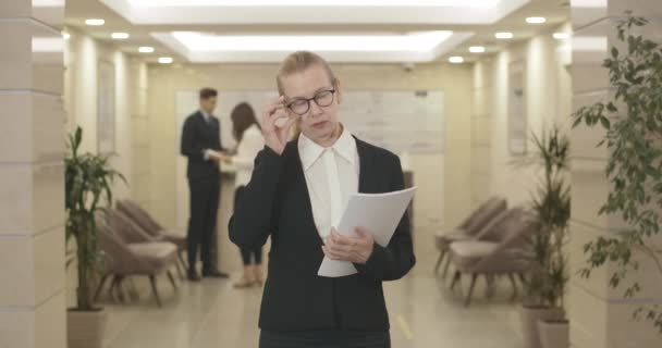 Portrait d'une femme d'affaires mi-adulte stricte en lunettes posant avec des documents dans le hall. Femme blanche professionnelle confiante debout dans le centre d'affaires et regardant la caméra. Siège social Cinema 4k ProRes. — Video